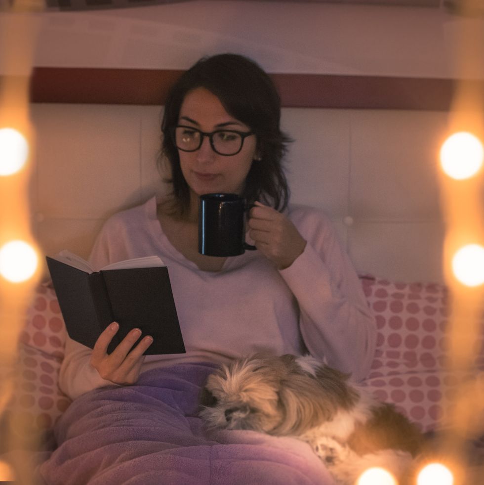 young woman holding mug reading book while sitting on bed at home