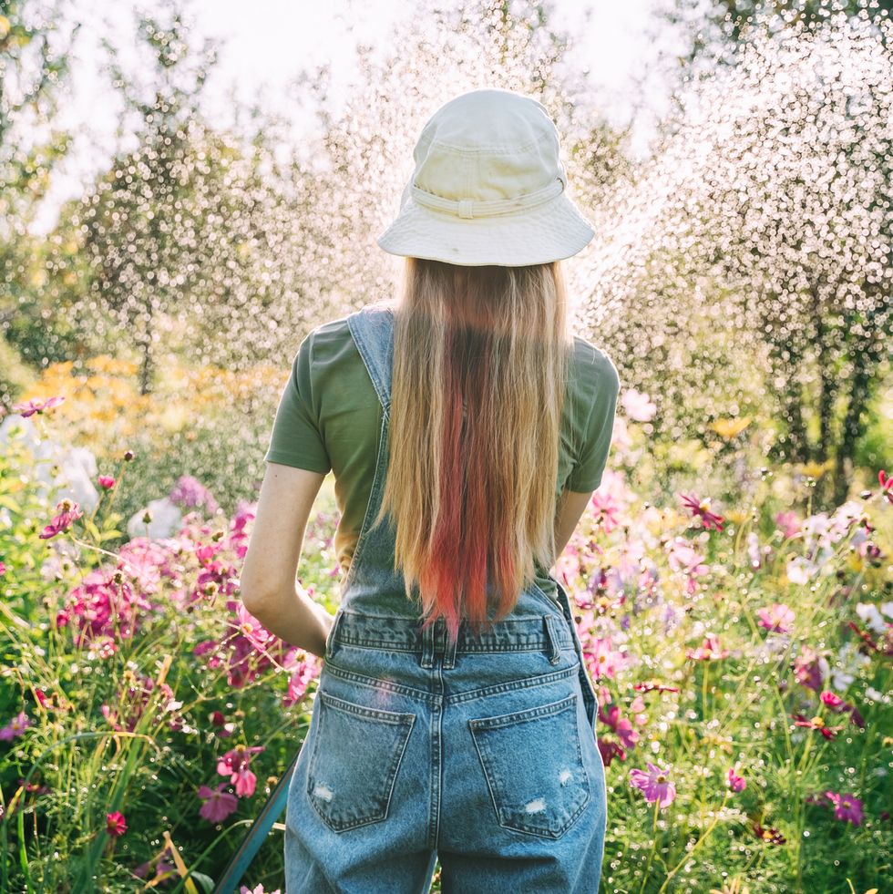 a young woman in a hat and denim overalls is watering flowers with a hose in the garden