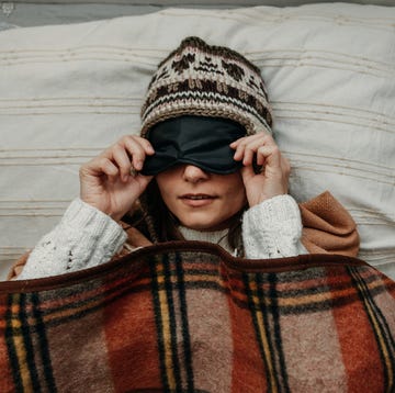 a young woman in a warm hat and sweater lifts a sleeping mask the girl is covered with a plaid blanket and lies in bed close up scandinavian style
