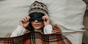 a young woman in a warm hat and sweater lifts a sleeping mask the girl is covered with a plaid blanket and lies in bed close up scandinavian style