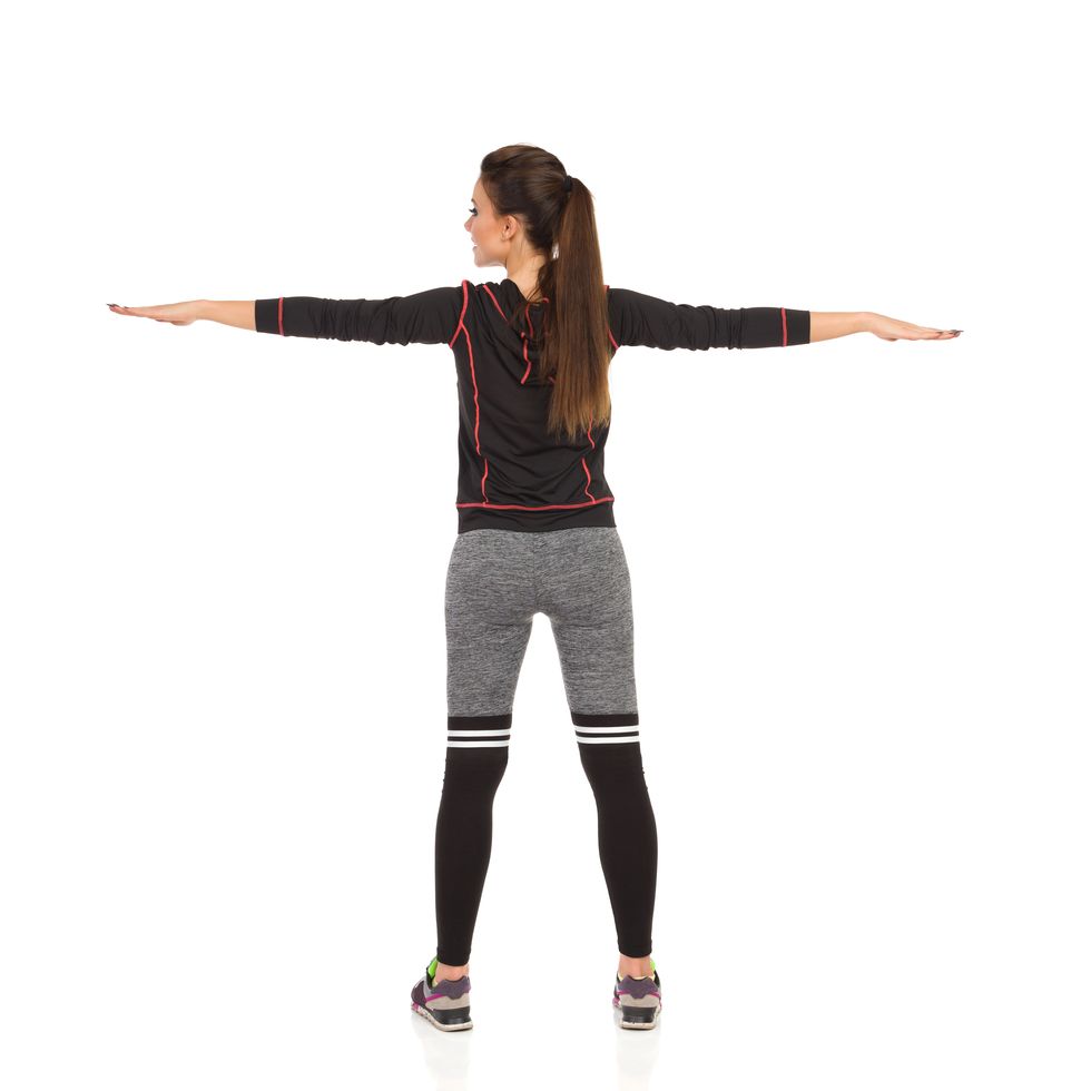 young woman in sports clothes is standing with arms outstretched rear view