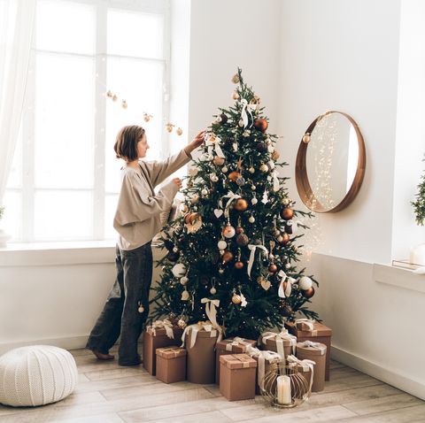 young woman is decorating christmas tree with many different decorations and festive garland in minimalist light interior new year celebration concept