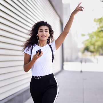 Young Woman Listening Music While Walking On Footpath By Wall