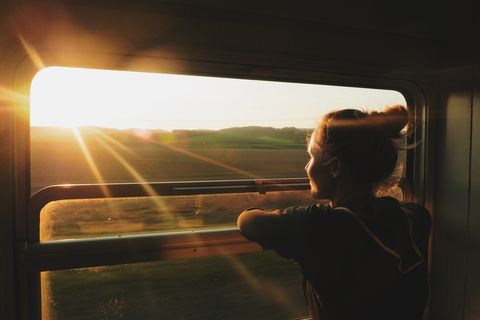 young woman looking through window while traveling in train during sunset
