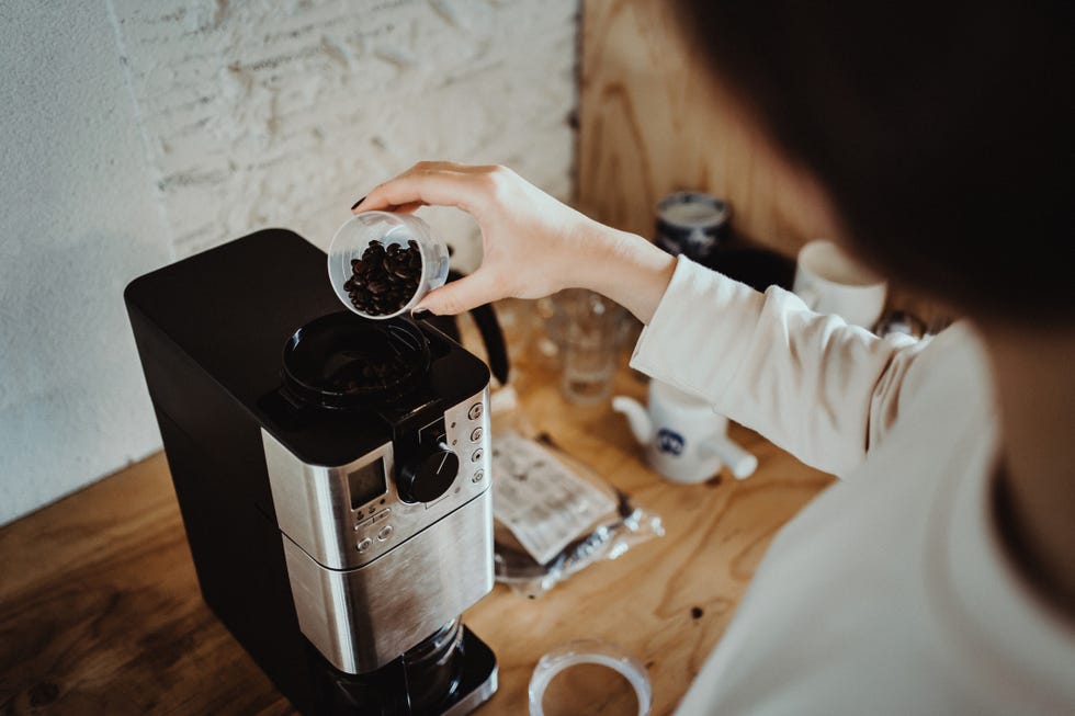 young woman measuring and pouring roasted coffee beans into coffee machine and preparing coffee at home in the early morning