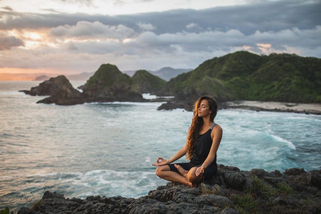 young woman practicing yoga in lotus pose at sunset with beautiful ocean and mountain view sensitivity to nature self analysis and soul searching spiritual and emotional concept