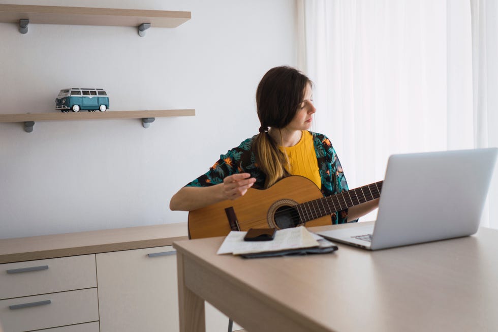 virtual happy hour ideas - Young woman sitting at table at home with laptop playing guitar