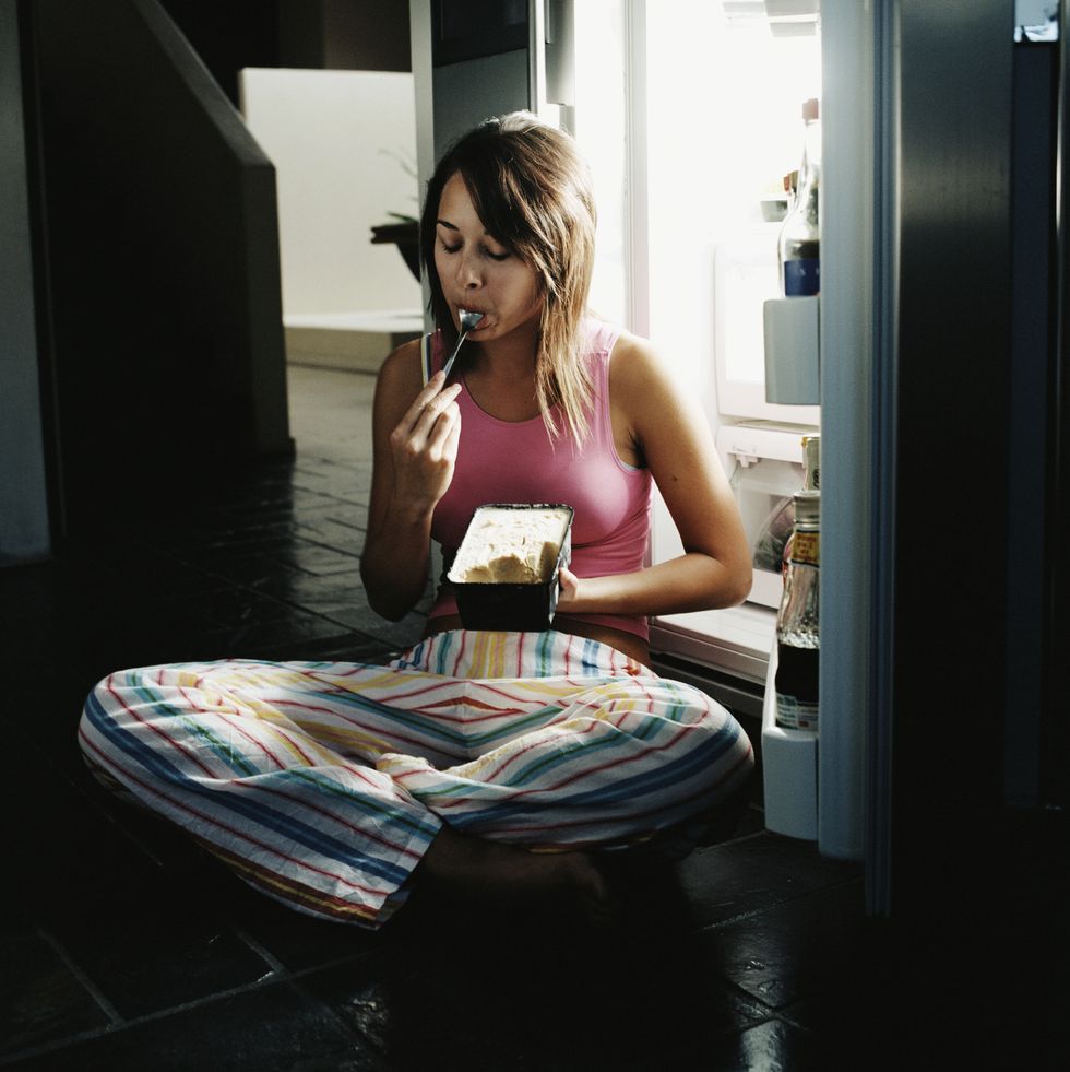 young woman sitting by open refrigerator, eating ice cream