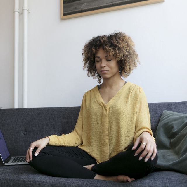 young woman sitting on couch at home next to laptop meditating
