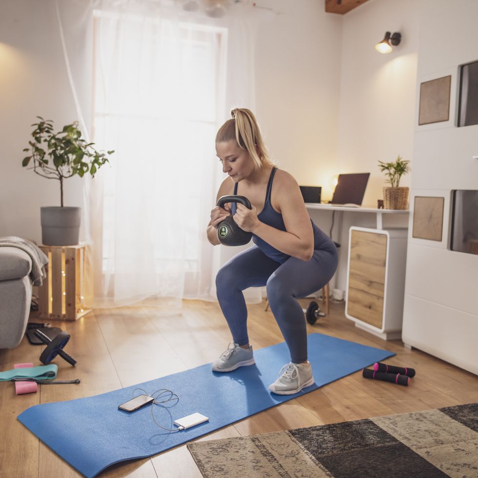 young woman squatting with kettlebell on yoga mat in living room