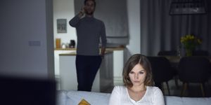 young woman sulking on sofa in evening, while boyfriend makes smartphone call
