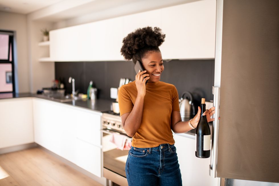 young woman taking a bottle of wine from the fridge and talking on the phone