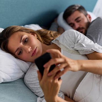 young woman using mobile phone and checking messages in bed while her husband is asleep