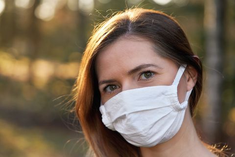 young woman wearing white cotton virus mouth nose mask, blurred sunset lit trees in background, closeup face portrait