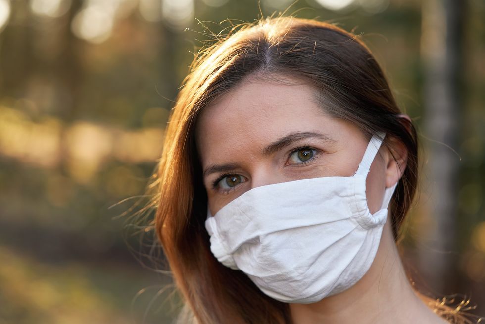 young woman wearing white cotton virus mouth nose mask, blurred sunset lit trees in background, closeup face portrait