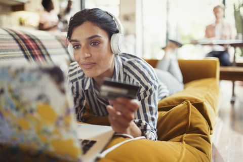 Young woman with credit card and headphones online shopping at laptop on living room sofa
