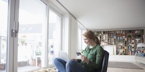 young woman with credit card and laptop sitting on a leather armchair in her living room
