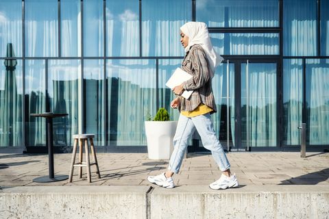 young  woman with hijab walking with laptop in front of glass facade