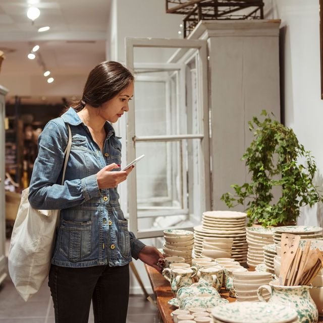 young woman with mobile phone choosing crockery arranged on table in boutique