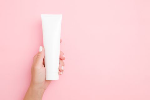 young woman's hand holding and showing white tube on light pink table pastel color care about clean and soft body skin daily beauty product close up empty place for text or logo top view