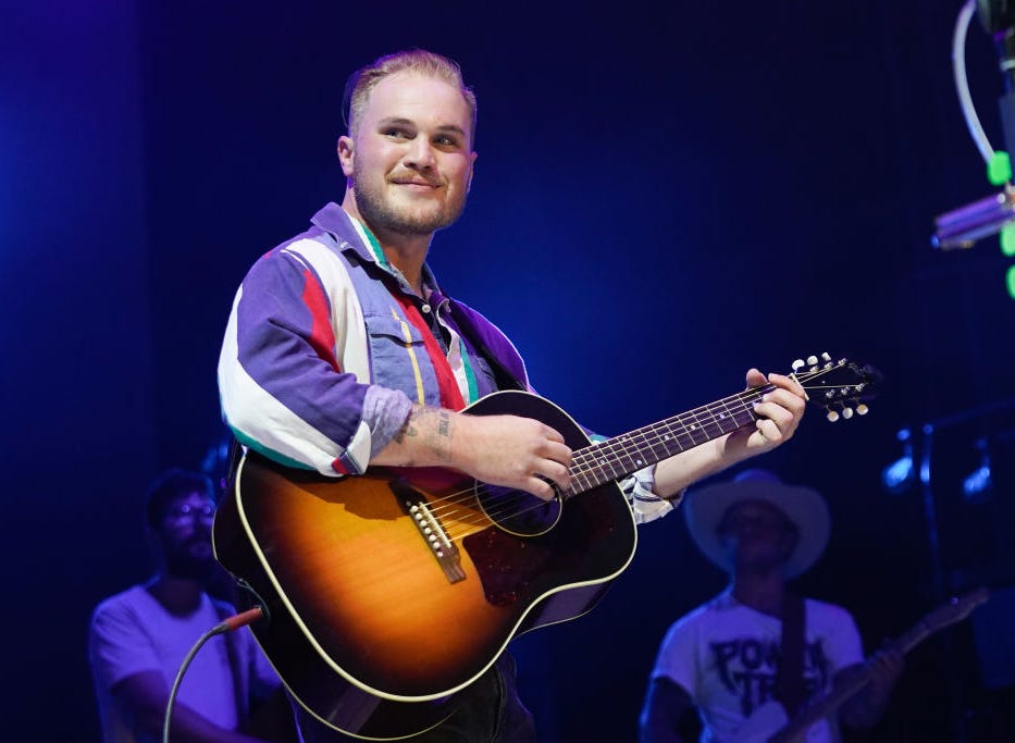 zach bryan looking out to his right into a crowd at a concert as he plays guitar on stage