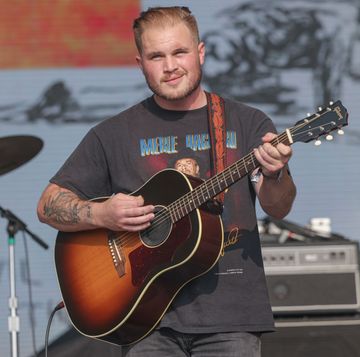 zach bryan strums a guitar as he stands on a stage, he wears a gray graphic t shirt with a slight smile on his face