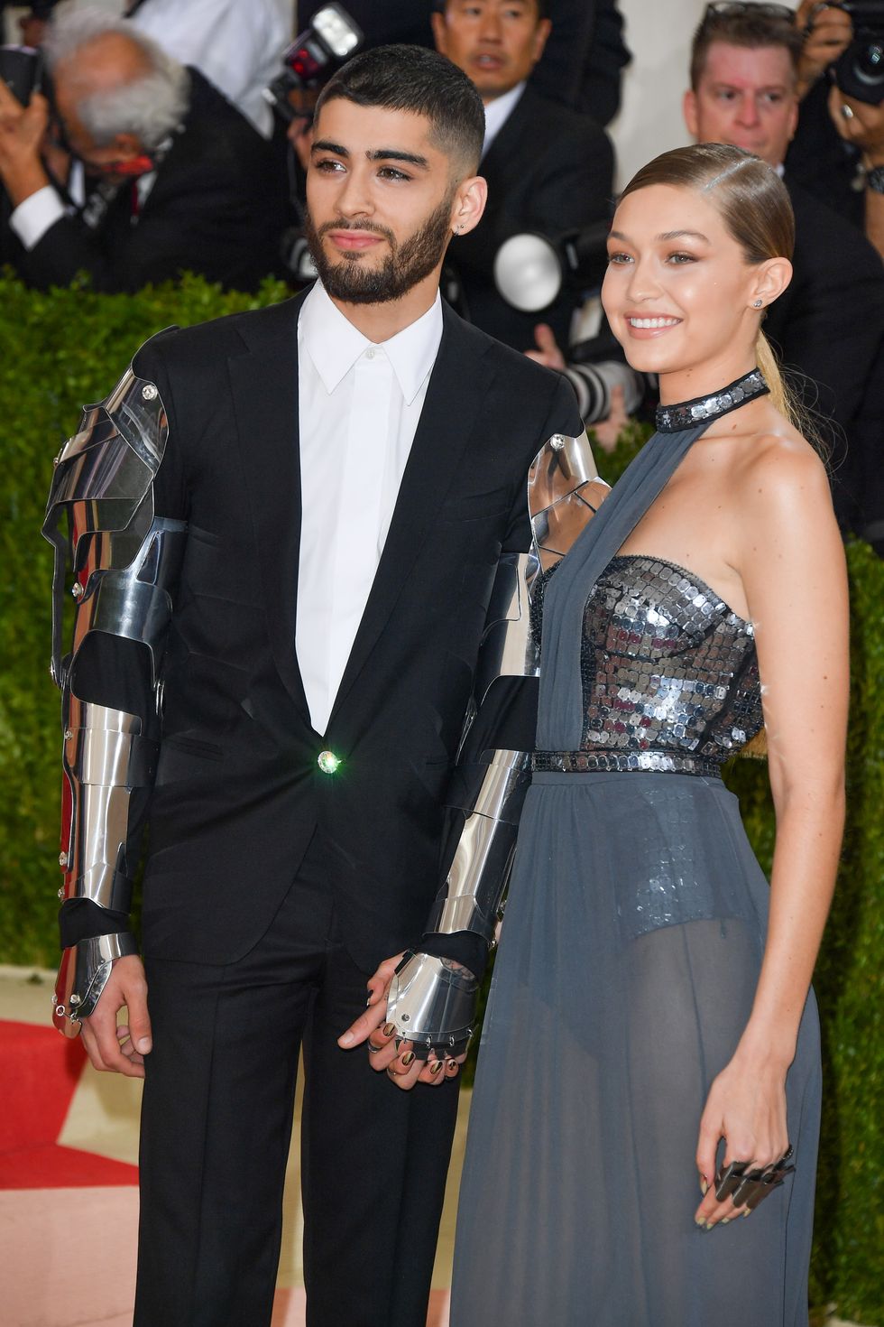 a man in a black suit white shirt and metal armor on his sleeves and a woman in a gray and silver gown hold hands and smiles for photos