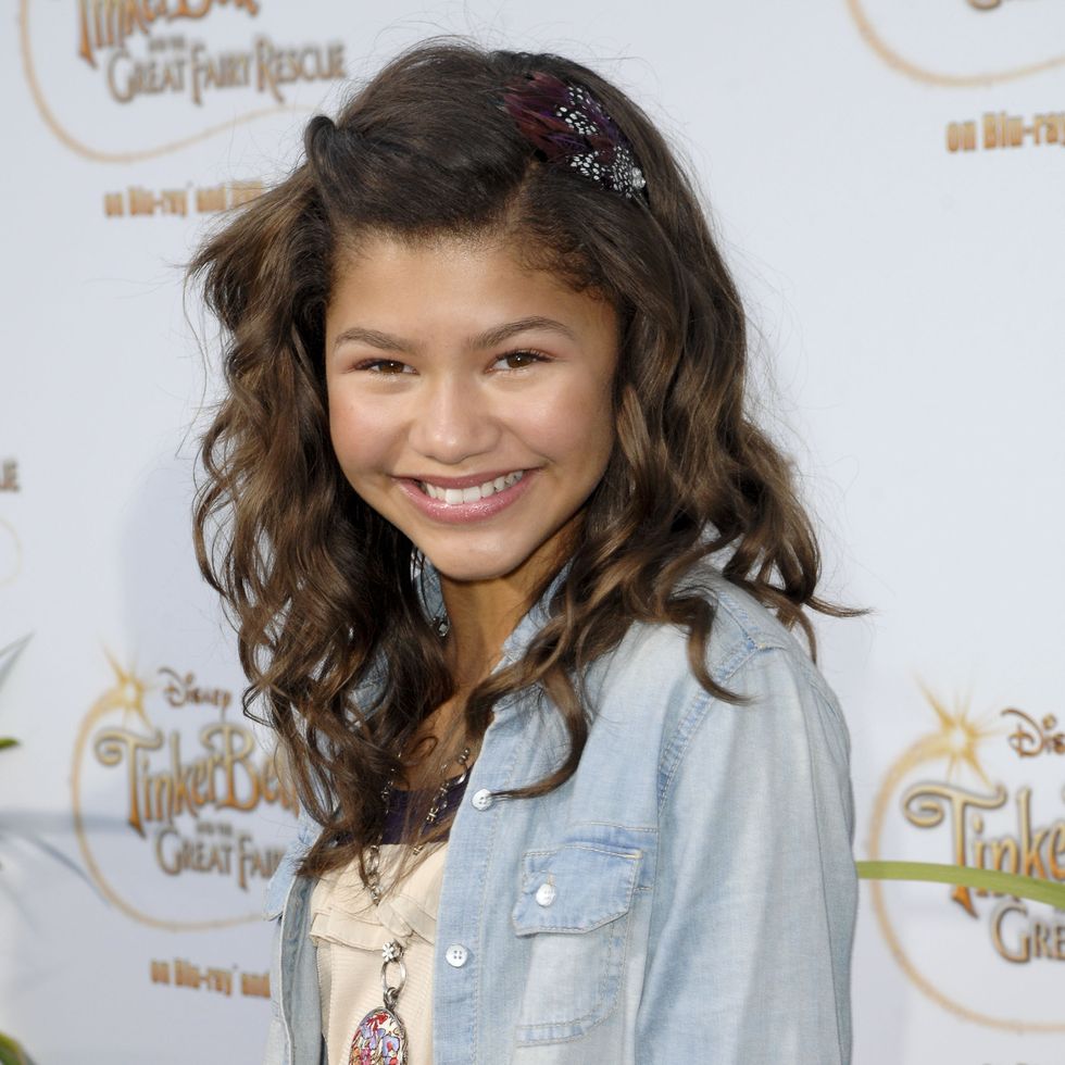 zendaya smiles at the camera, she wears a denim collared shirt over a light colored dress with a long necklace and a feather in her hair