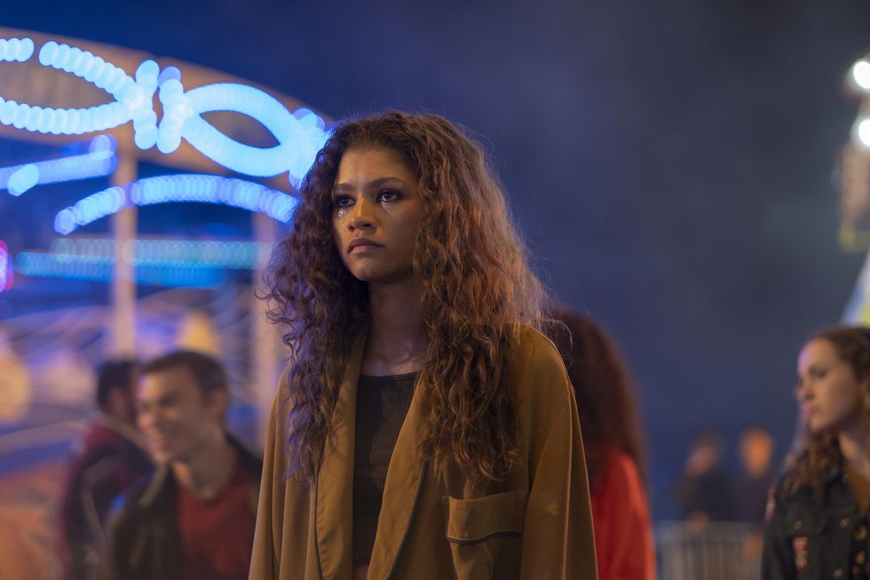 zendaya stands outside and looks to the left, she wears a brown jacket over a dark colored shirt