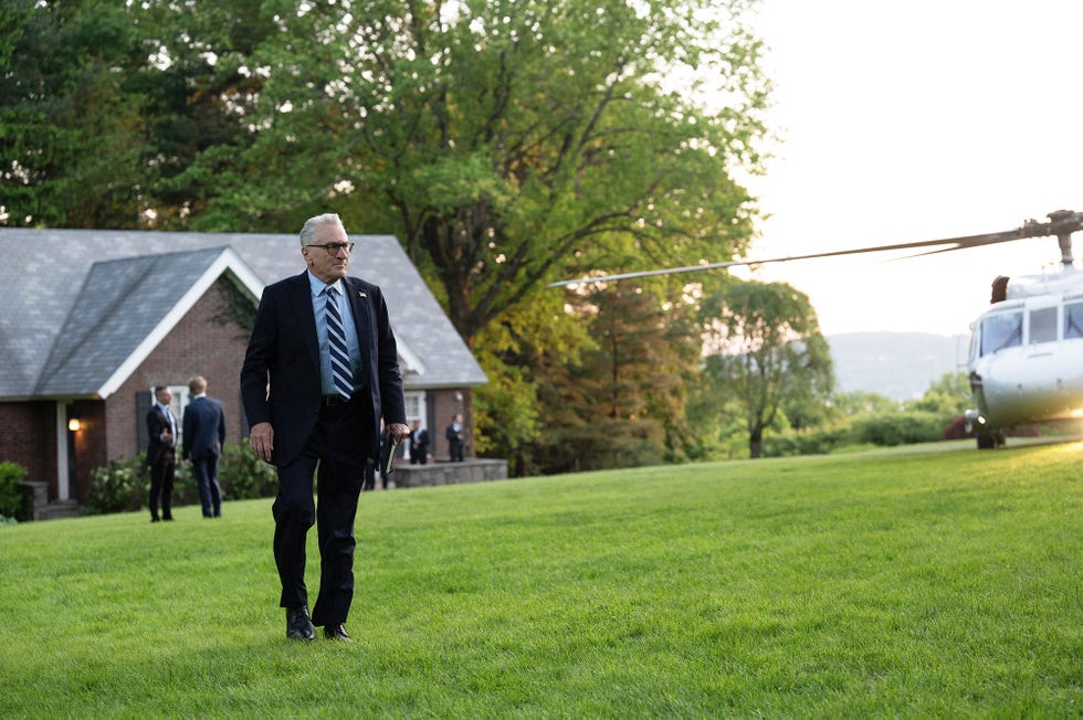 man in a suit walking across a lawn near a helicopter and a house