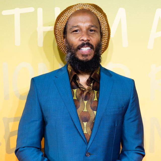 ziggy marley smiles while standing in front of a green, yellow and orange photo background, he wears a blue suit with a green and brown patterned shirt and brown knit cap