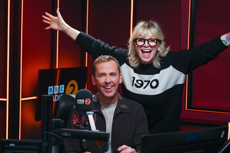 zoe ball raises her arms with joy as she stands in the radio 2 breakfast show studio with her friend and successor scott mills