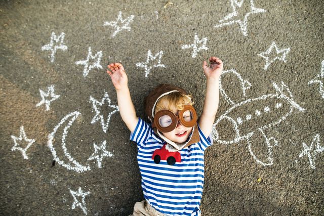 Portrait of smiling toddler wearing pilot hat and goggles lying on asphalt painted with airplane, moon and stars