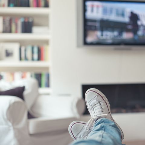 Picture of jeans and sneakers, feet up on couch, with television, fireplace, sofa and bookcase in background.