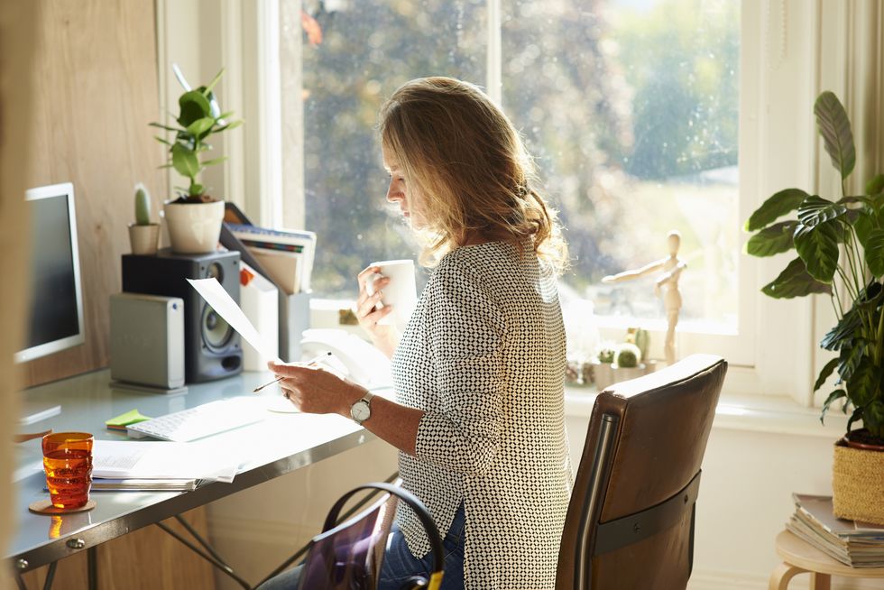 Woman drinking coffee and reading paperwork at desk in sunny home office.