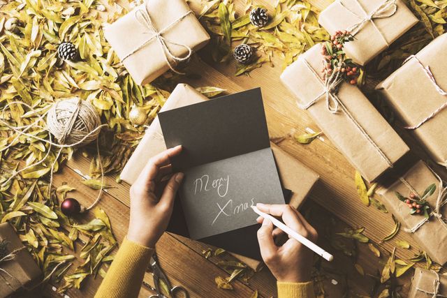 Christmas concept table top flat lay on wooden background. Woman wearing yellow sweater writing Merry Xmas on grey greeting card. Jute string, gift boxes, Christmas ornament, pine cones, firethorn branches and yellowed leaves on wooden background.