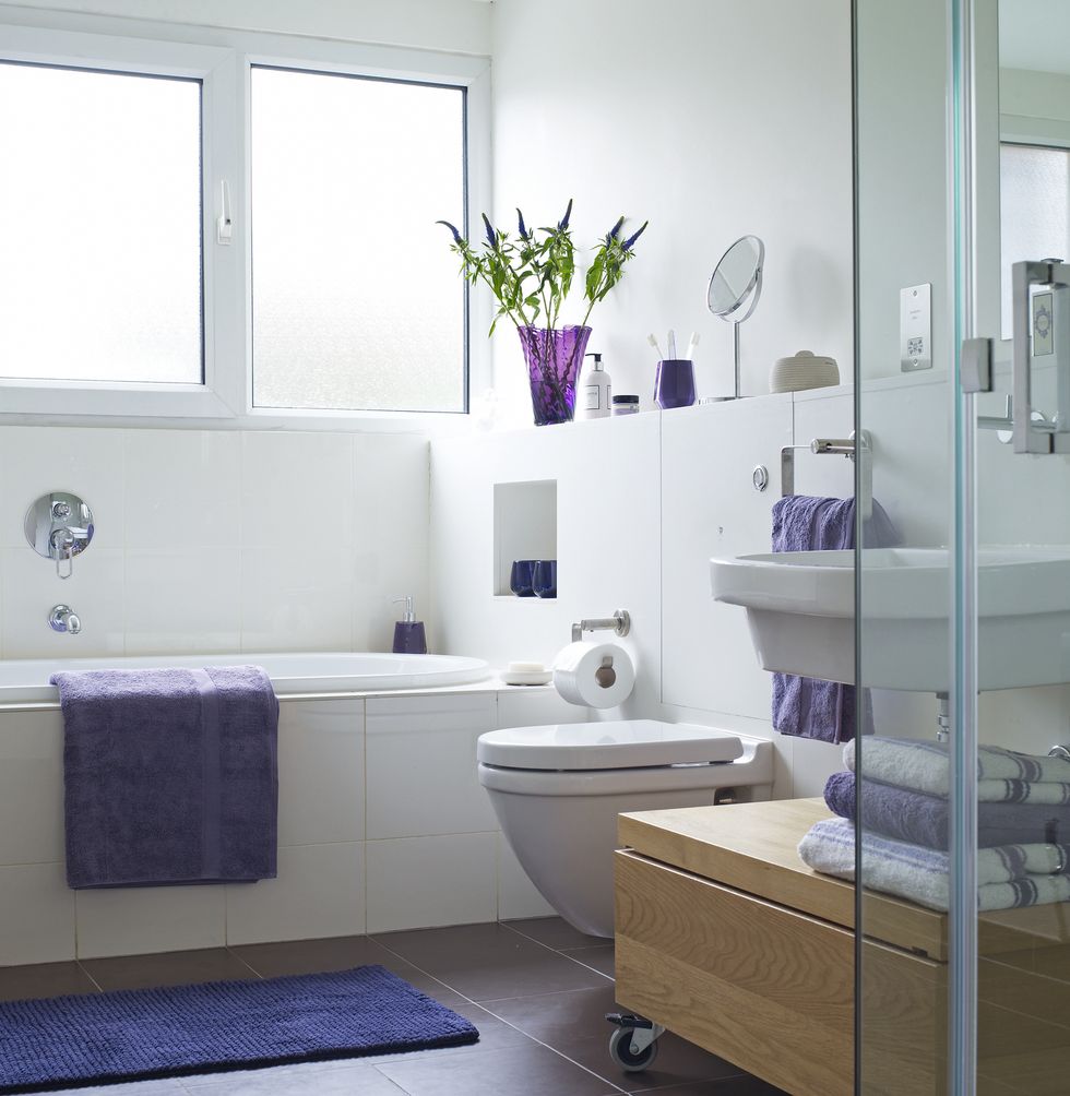 Brightly lit bathroom with purple towel on side of bath and folded towels near shower