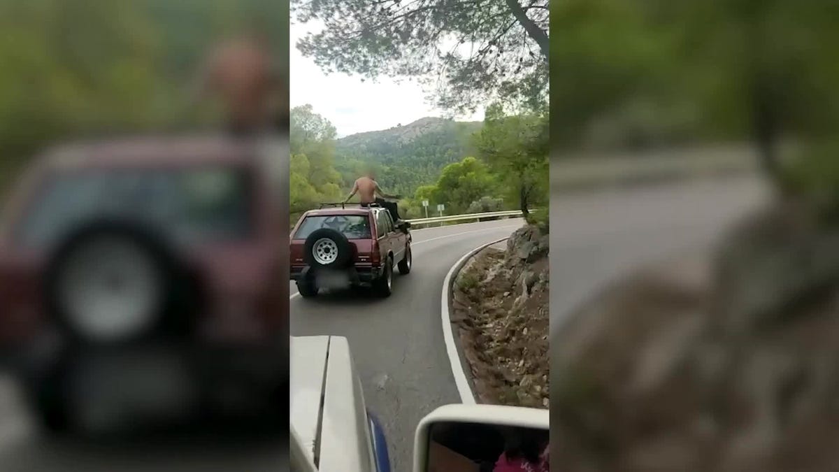 He descends a mountain pass on the roof of a car, shirtless… while being filmed