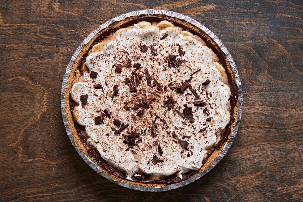 banoffee pie topped with whipped cream and shaved chocolate in a foil pie tin on a dark brown background