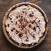 banoffee pie topped with whipped cream and shaved chocolate in a foil pie tin on a dark brown background