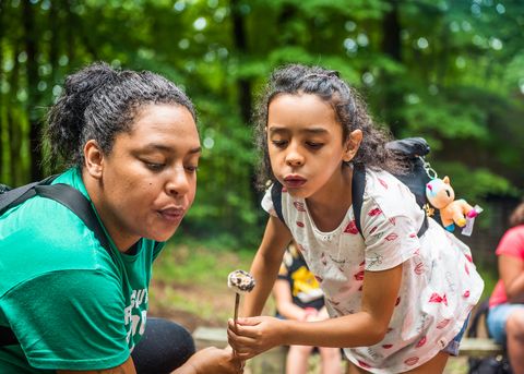 A Girl Scout of the USA  and her troop leader blow on a roasted marshmallow at a campout.